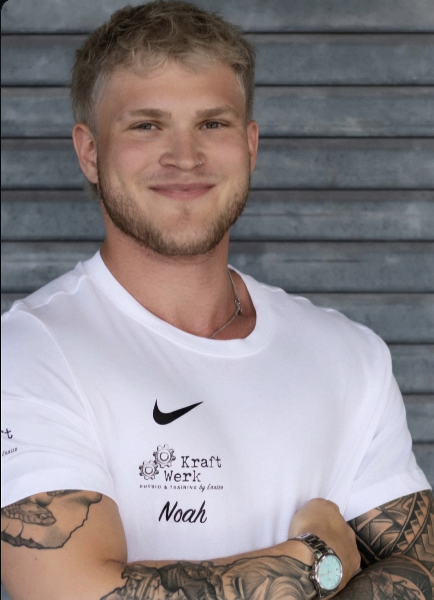 Junger Mann mit blonden Haaren und Tattoos lächelt selbstbewusst in weißem Shirt